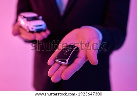 Cropped photo of a young attractive businessman in a black suit and white shirt holding a model car in the palm of his hand, isolated on a neon pink background. Car purchase, rental.