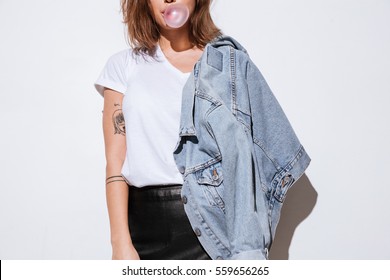 Cropped photo of young lady dressed in jeans jacket standing isolated over white background while blowing bubble with chewing gum.