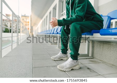 Cropped photo of a young man's legs in a green suit and bloodsuckers, sitting alone in the stands and in pain. Background. Fashionable street photo of a male model in a green sports suit.