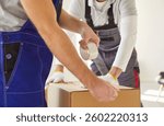 Cropped photo of young men workers packing cardboard box with adhesive tape planning to move to new apartment. Movers male team of moving service working in overalls. Relocation concept