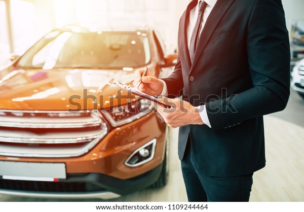 Cropped photo of young salesman in full suit\
standing on dealership background while paperwork with documents in\
hands