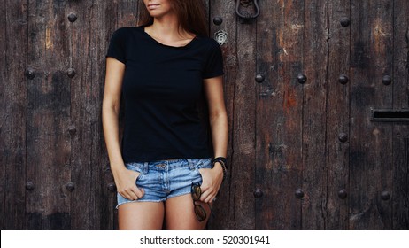 A cropped photo of a young woman wearing black blank t-shirt and blue jeans shorts standing on the wooden door background. Empty space for text or design.
