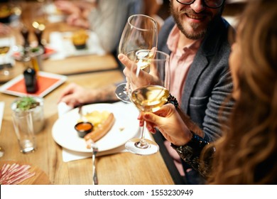 Cropped picture of cute caucasian couple sitting in restaurant and cheering with wine. In background are their friends.