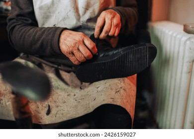 Cropped picture of an old craftsman holding a boot in his hands and fixing a zipper. An unrecognizable cobbler is sitting at his workshop in his apron with a boot in his hands. Hands fixing zipper. - Powered by Shutterstock