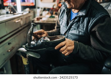 Cropped picture of a senior cobbler sitting at workshop and using hammer for fixing a leather shoe. A senior shoemaker is running small business workshop. A shoemaker is repairing a leather shoe. - Powered by Shutterstock