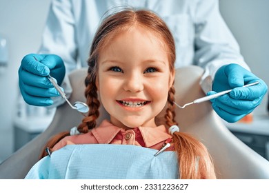 Cropped portrait of little girl with pigtails hair sitting in dental chair looking at camera and smiling. Behind, a doctor in gloves holds examination tools.Children's dentistry. - Powered by Shutterstock