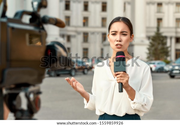 Cropped
portrait of professional female reporter at work. Young woman
standing on the street with a microphone in hand and smiling at
camera. Horizontal shot. Selective focus on
woman