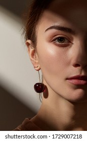 Cropped Portrait Shot Of A Young European Woman With Picked Up Brown Hair. The Lady Is Wearing A Stud Earring Made As A Red Translucent Cherry With A Golden Tail.