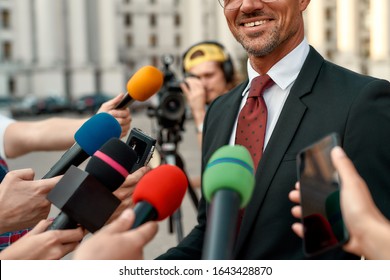 Cropped Portrait Of Smiling Man In Suit Giving Interview To Reporters. People Making Interview Using Microphones And Equipment Set At Outdoor Location. Horizontal Shot. Selective Focus On Man