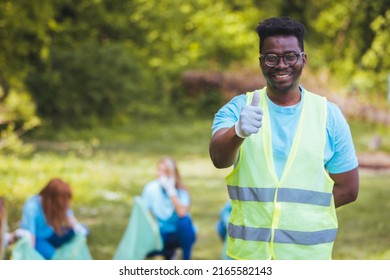 Cropped Portrait Of A Young African Male Volunteer Doing Community Service In The Local Park With His Friends In The Background. Thumb Up