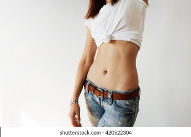 Cropped portrait of young female with flat belly and slim waist standing on blank studio wall.  Brunette woman wearing denim pants showing her beautiful fit body. Lifestyle and people concept. 