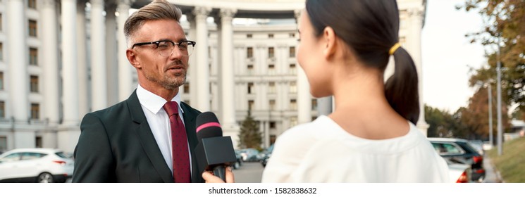 Cropped Portrait Of Young Woman Conducting Journalistic Interview Of Cheerful Politician. People Making Interview Using Equipment Set At Outdoor Location. Horizontal Shot. Selective Focus On Man