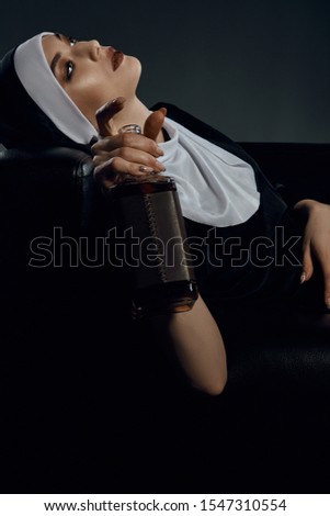 Cropped profile shot of a nun, sitting back in a chair. She's holding bottle of wine in her hand and looking forward. The nun is wearing dark nun's clothes. 