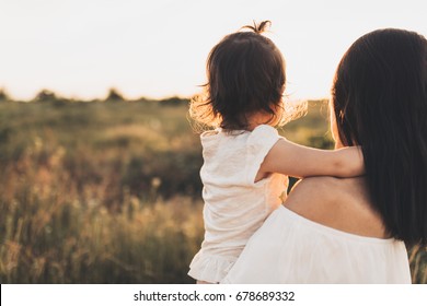 Cropped rear view of Caucasian brunette mother and child daughter embrace each other on meadow sunset. Beautiful brunette woman hugs her toddler girl and looking on sunset background. Motherhood. - Shutterstock ID 678689332