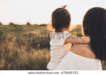 Cropped rear view of Caucasian brunette mother and child daughter embrace each other on meadow sunset. Beautiful brunette woman hugs her toddler girl and looking on sunset background. Motherhood.