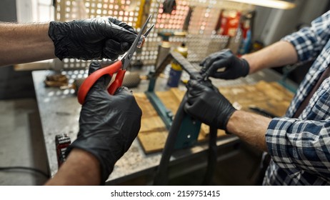 Cropped Selective Focus Of Cycling Mechanic Hands In Gloves Cutting Patch With Scissors For His Blurred Colleague Glueing Bicycle Tube In Workshop. Teamwork. Bike Service, Repair And Upgrade