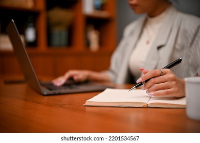 Cropped And Selective Focus Image, A Professional Asian Female Boss Or Businesswoman Working At Her Office Desk, Using Laptop And Writing Something On Notebook.