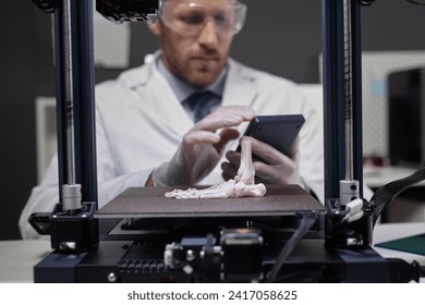 Cropped shot of 3D printer frame with focus on human foot bone model on hot bed and male engineer working with device controller in background - Powered by Shutterstock