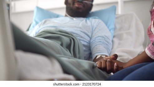 Cropped shot of african wife visiting husband in hospital holding hands and supporting. Sick young black male patient lying in hospital bed and talking to woman visitor in ward - Shutterstock ID 2123170724