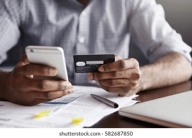 Cropped Shot Of African-American Male Paying Bill At Restaurant With Online Payment Technology Via Internet, Using Free Wi-fi During Breakfast, Sitting At Table With Plastic Card And Smart Phone