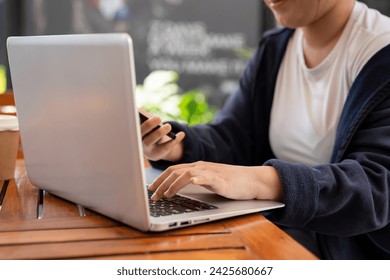 A cropped shot of an Asian woman multitasking, using her smartphone and typing on a laptop computer, while working remotely at a cafe in the city.