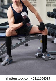 Cropped Shot Of An Athletic Man In A Sportswear And Black Knee Socks With A Zipper. He Is Sitting On A Bench With A Dumbbell In His Hand. The Man In Socks Is Isolated On The Gym Background.  