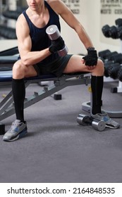 Cropped Shot Of An Athletic Man In A Sportswear And Black Knee Socks With A Zipper. He Is Sitting On A Bench With A Dumbbell In His Hand. The Man In Socks Is Isolated On The Gym Background.           