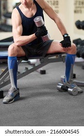 Cropped Shot Of An Athletic Man In A Sportswear And Blue Knee Socks With A Color Print. He Is Sitting Down On A Bench With A Dumbbell In His Hand. The Man In Golfs Is Isolated On The Gym Backgroun