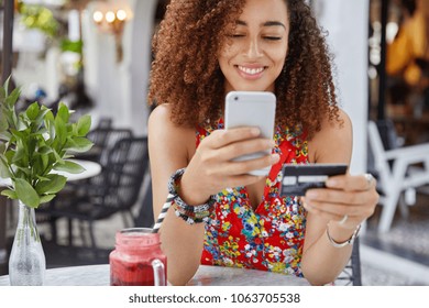Cropped shot of beautiful African American woman with happy look, holds modern cell phone and credit card, makes shopping online, drinks smoothie in outdoor cafeteria. People and payment concept