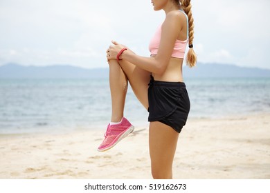 Cropped shot of blonde Caucasian girl with beautiful fit body doing stretching exercises before jogging workout, standing on beach against blurred ocean background. Sporty woman warming up outdoors