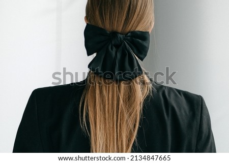 Cropped shot of a blonde woman with long hair in a black jacket. Her hair is tied with a hairpin adorned with a black bow. The photo was taken against a blue background.
