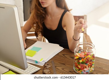 Cropped shot of the Caucasian female office worker taking marmalade from the big jar at work. The girl eats candies for improvement of work of a brain during hard work over the financial statement