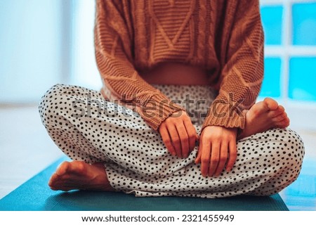 Cropped shot of a caucasian girl in a dotty yoga pants sitting on a mat in a lotus pose with her hands crossed forward. Wearing a knitted sweater and long legged pants while enjoying her morning yoga.