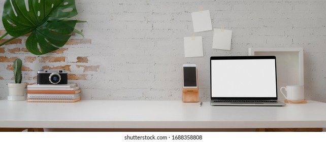gepfropfte Aufnahme von bequemem Arbeitsplatz mit kleinem Laptop, Smartphone, Kameradekoration und Kopienraum auf weißem Tisch mit Ziegelwand 