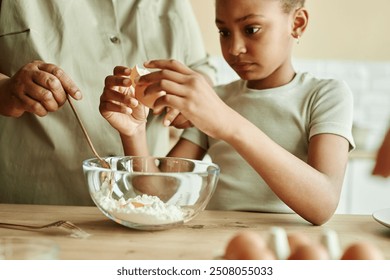 Cropped shot of concentrated African American little girl carefully cracking egg in bowl at wooden counter, learning how to cook pancakes together with granny at beige kitchen - Powered by Shutterstock