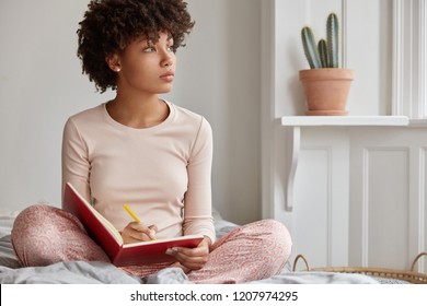 Cropped shot of dark skinned woman sits crossed legs, wears pyjamas, makes notes in diary, focused aside, blank copy space against domestic interior. Blogger creats publication for blog in notebook - Shutterstock ID 1207974295