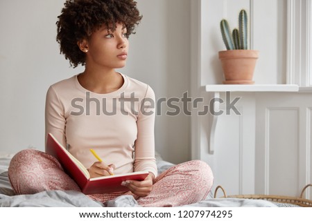 Cropped shot of dark skinned woman sits crossed legs, wears pyjamas, makes notes in diary, focused aside, blank copy space against domestic interior. Blogger creats publication for blog in notebook
