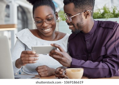 Cropped Shot Of Deighted Happy African Couple Holds Smart Phone Horizontally, Watch Interesting Video, Have Coffee Break, Smile Joyfully, Wears Round Spectacles. Dark Skinned Woman And Man Friends