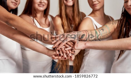 Cropped shot of diverse women in white shirts showing unity, putting hands on top of each other like a team while standing isolated over grey background. Selective focus on hands. Web Banner