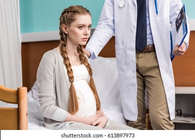 Cropped Shot Of Doctor Standing Near Sad Pregnant Woman Sitting On Hospital Bed 