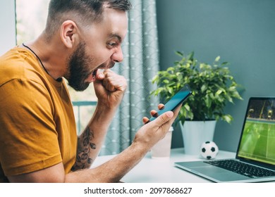 Cropped Shot Of Euphoric Soccer Fan Man Celebrating Moneu Win After Betting At Favourite Team Using Bookmaker's Mobile Application. Young Guy Watching Football Play Live Broadcast On His Laptop.