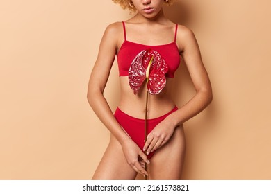 Cropped shot of faceless young woman holds calla flower over body wears red top and panties poses over beige background demonstrates natural beauty and skin imperfections. Body care concept.