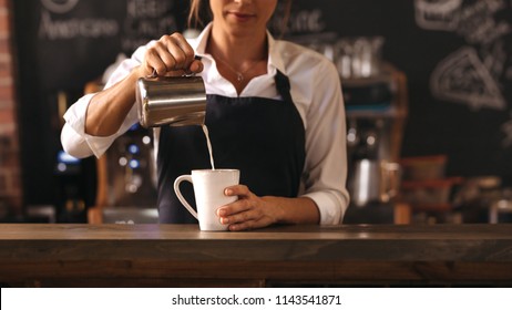 Cropped shot of female barista making a cup of coffee while standing behind cafe counter. Young woman pouring milk into a cup of coffee. - Shutterstock ID 1143541871