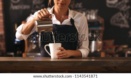 Cropped shot of female barista making a cup of coffee while standing behind cafe counter. Young woman pouring milk into a cup of coffee.