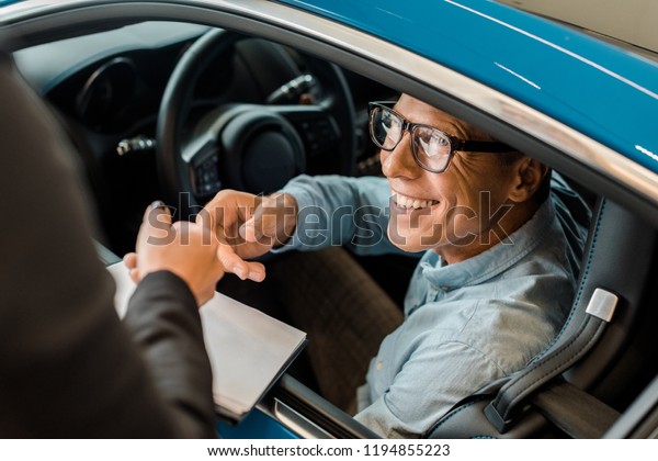 cropped shot of female car\
dealer passing car dealership contract to smiling adult man in\
showroom
