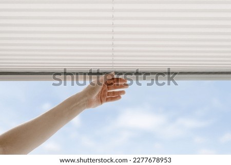 cropped shot of female hand close pleated jalousie on window in living room, controlling lighting range, protection of sunlight, blue sky view 