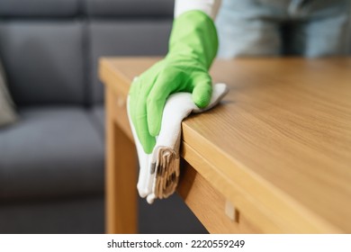 cropped shot of female hand in green rubber glove hold fabric rag and wipe wooden table from dust in living room, household chores concept