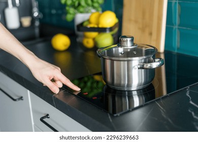 cropped shot of female hand press button on induction hob and choose temperature on control panel for cooking dinner in saucepan at home kitchen with stylish interior