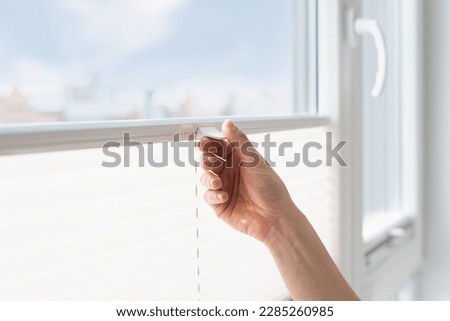 cropped shot of female hand pull up and close pleated jalousie on window in living room, controlling lighting range, protection of sunlight, blue sky view 