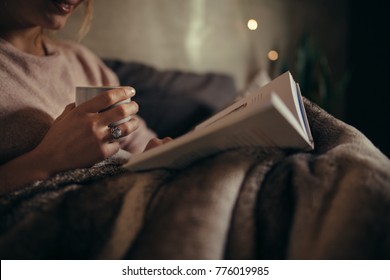 Cropped shot of female hands with book and coffee. Female reading book on bed at night.